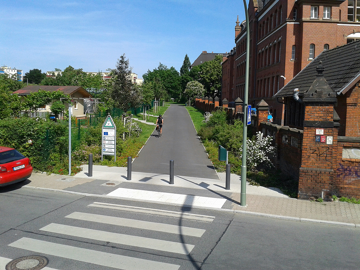 Blick auf Zebrastreifen, links Gärten, rechts Klinkerbauten, dazwischen breiter Radweg mit Radfahrer