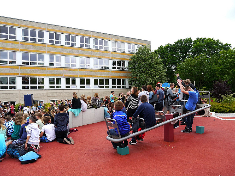 roter Kunststoffboden, Schüler von hinten auf Wippe und Sitzstufen, im Hintergrund Schule