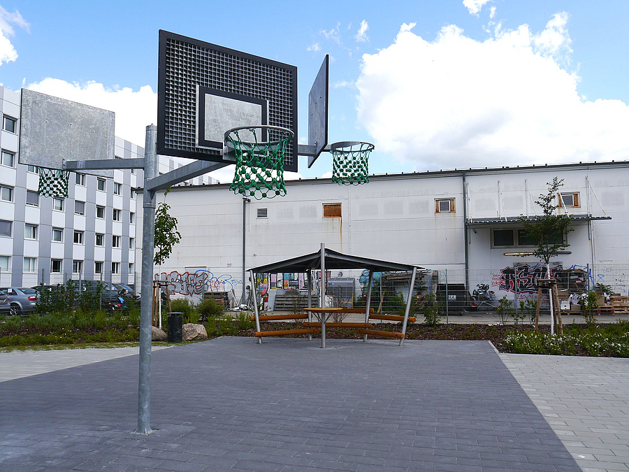Mast mit drei Basketballkörben auf Pflasterfläche, dahinter Pavillon vor Flachbau