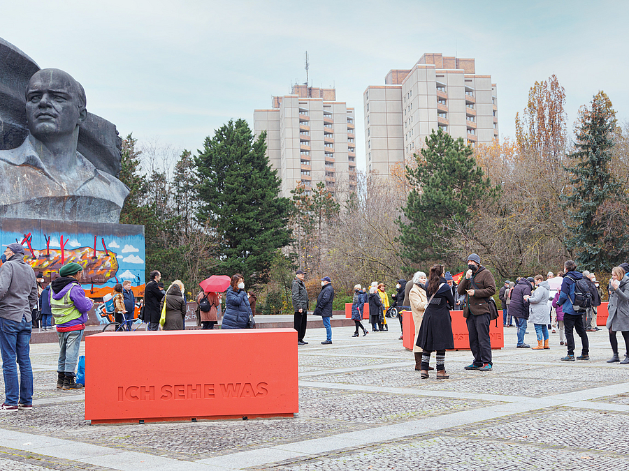 Monumentales Denkmal, roter Quader auf Platz mit vielen Menschen, dahinter Hochhäuser