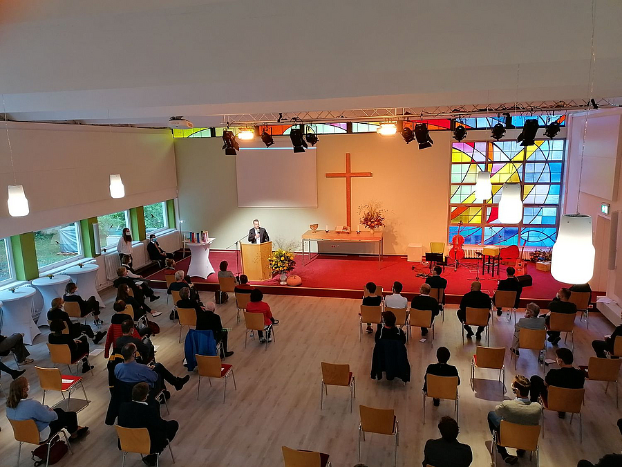 Saal mit farbigem Glasbildfenster und Kreuz an der Wand, Publikum mit Abstand