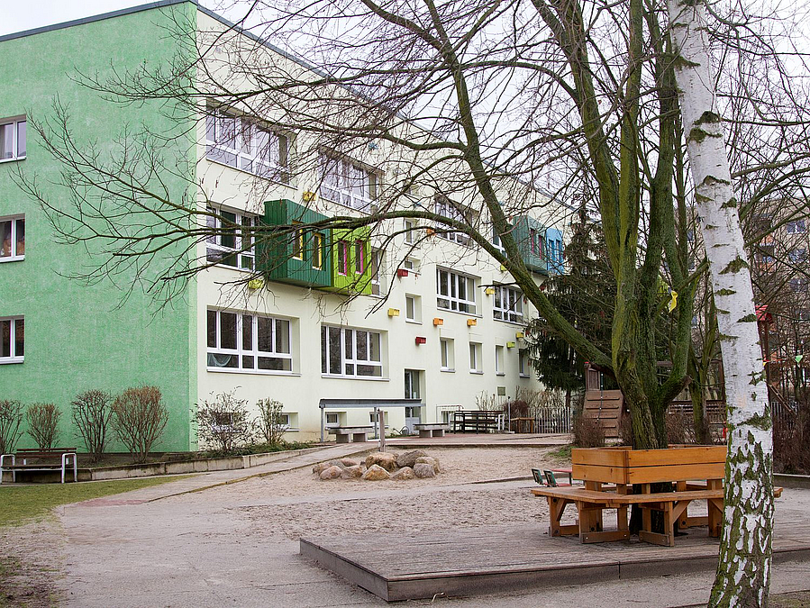 Blick über wintelich kahle Bäume und Spielgeräte auf Dreigeschosser mit grünem Giebel und heller, in Grüntönen abgesetzter Fassade
