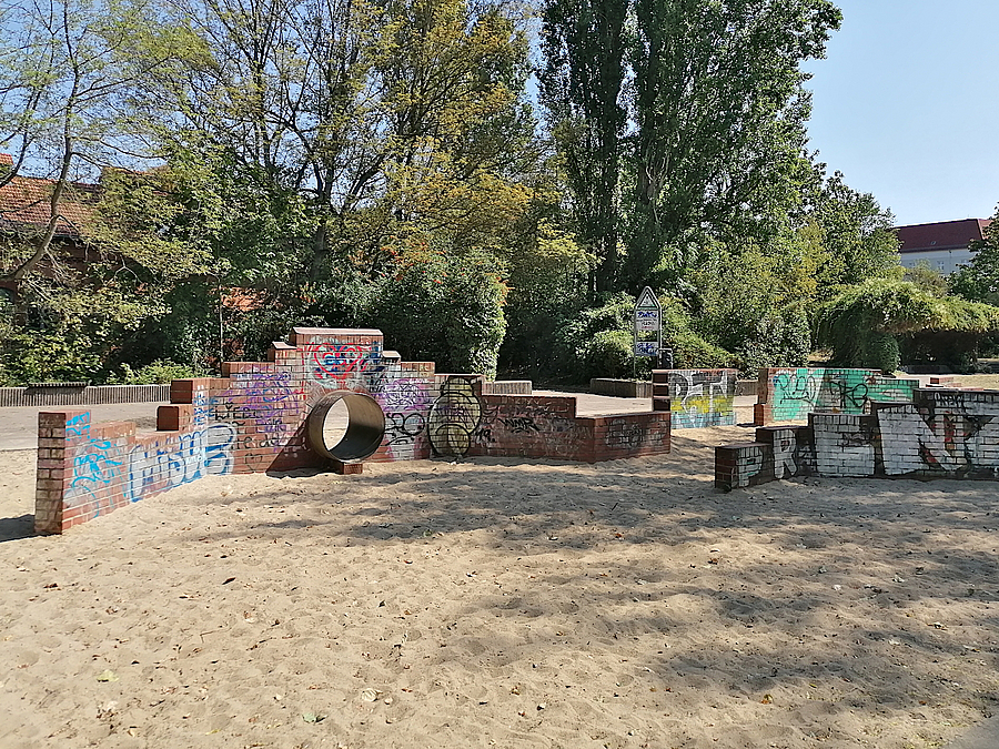 Abgestufte, niedrige Klinkermauer mit Graffiti auf Sandfläche, dahinter Bäume