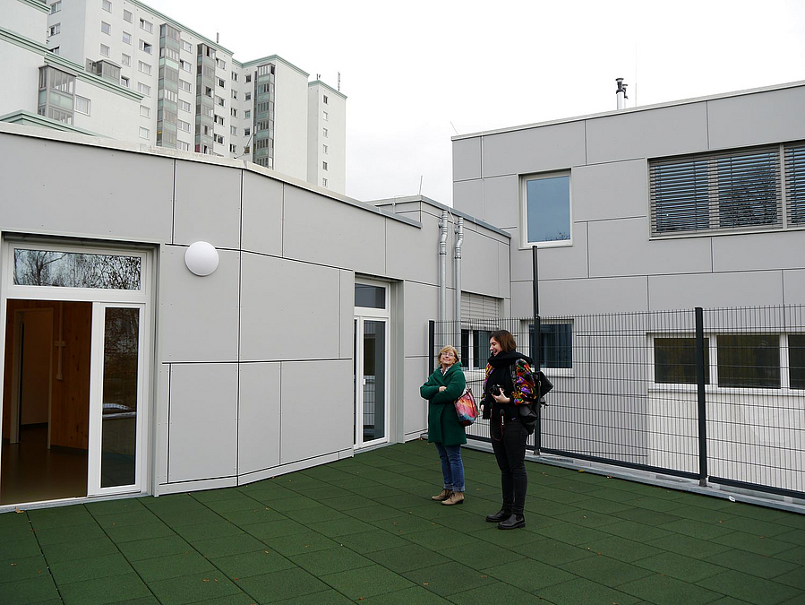 Gebäudewinkel zwischen unterschiedlich hohen Teilen in Grau, grüner Bodenbelag