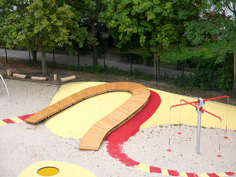 rot-gelber Kunststoffpfad auf Sandfläche zu gelbem Kunststoffhügel mit Holzweg
