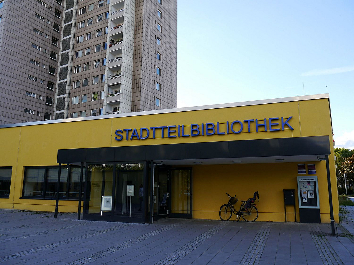 Gelber Flachbau mit Vordach und Schriftzug "Stadtteilbibliothek", Fahrrad, im Hintergrund Hochhaus