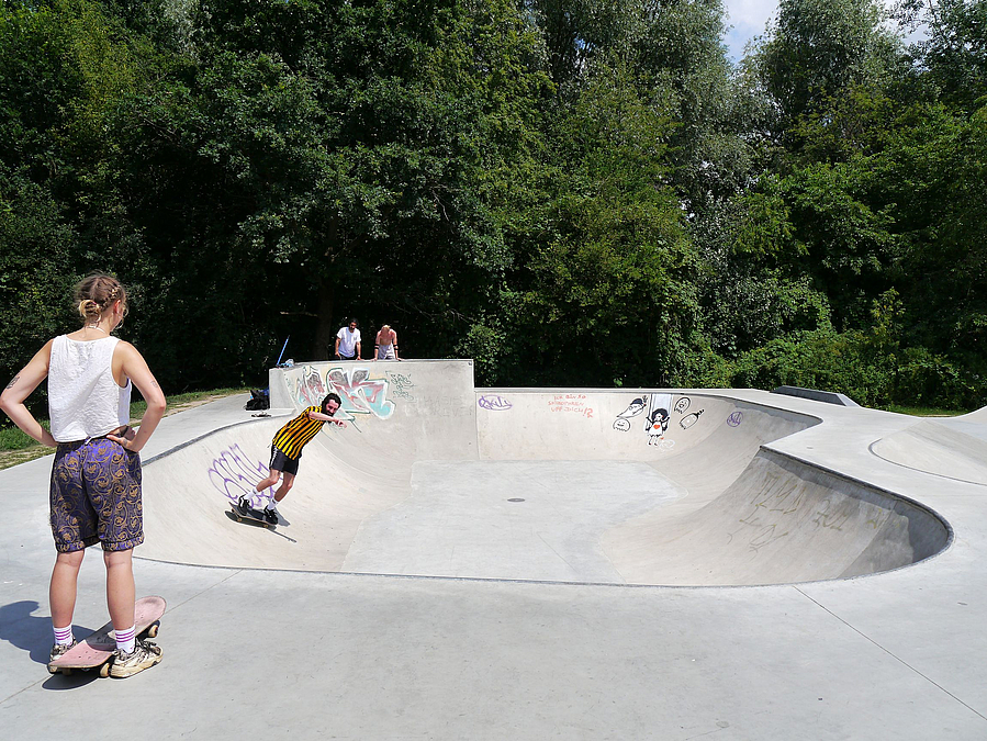 Bowl, junge Frau am Rand mit Skateboard und Skater innen