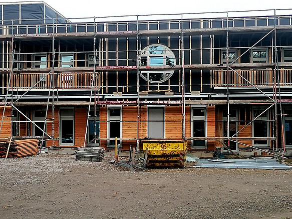 Baugerüst vor zweistöckiger Fassade, unten Holz, oben schwarz mit rundem Fenster im OG