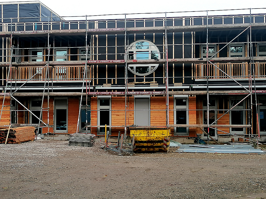 Baugerüst vor zweistöckiger Fassade, unten Holz, oben schwarz mit rundem Fenster im OG