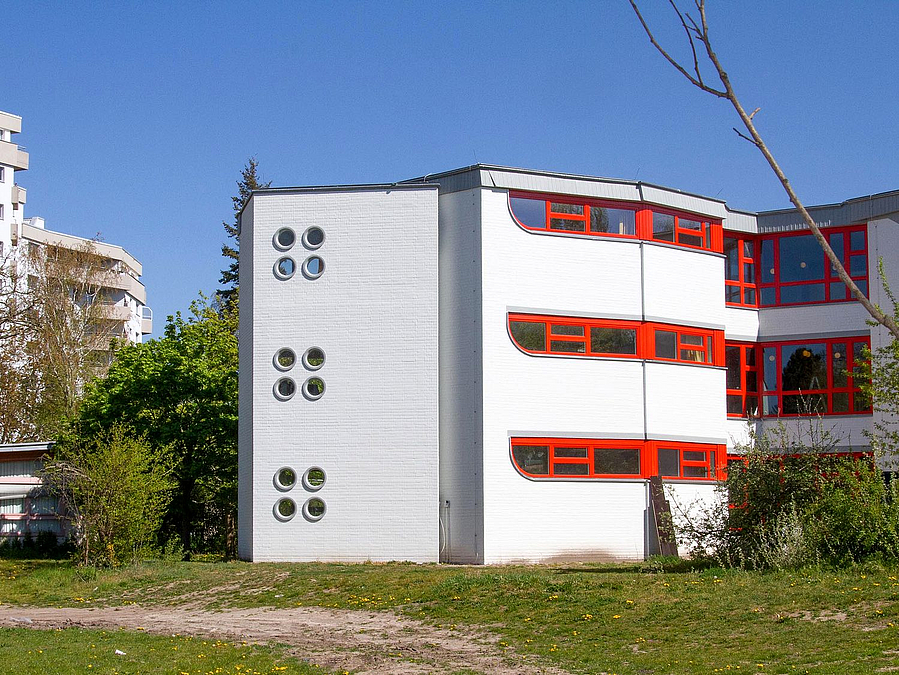 Dreistöckiges, weißes Gebäude mit schmalen roten Fensterbändern und Treppenturm mit Bullaugen