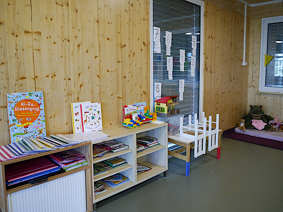 Raum mit Wänden aus hellem Holz, niedrige Regale mit Kinderbüchern