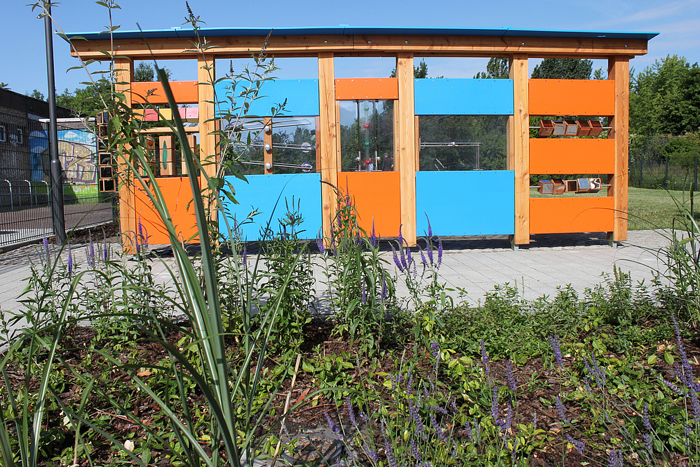 Mannshoher, blau-Oranger Rahmen mit Fenstern, in denen u.a. eine Kugelbahn zu sehen ist, davor Wiesenblumen