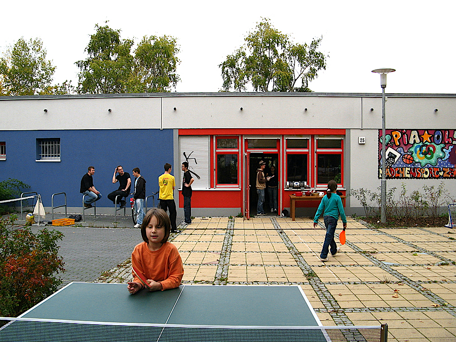 Eingeschossiger Bau in weiß und blau mit rotem Türrahmen, davor Tischtennisplatte mit Mädchen