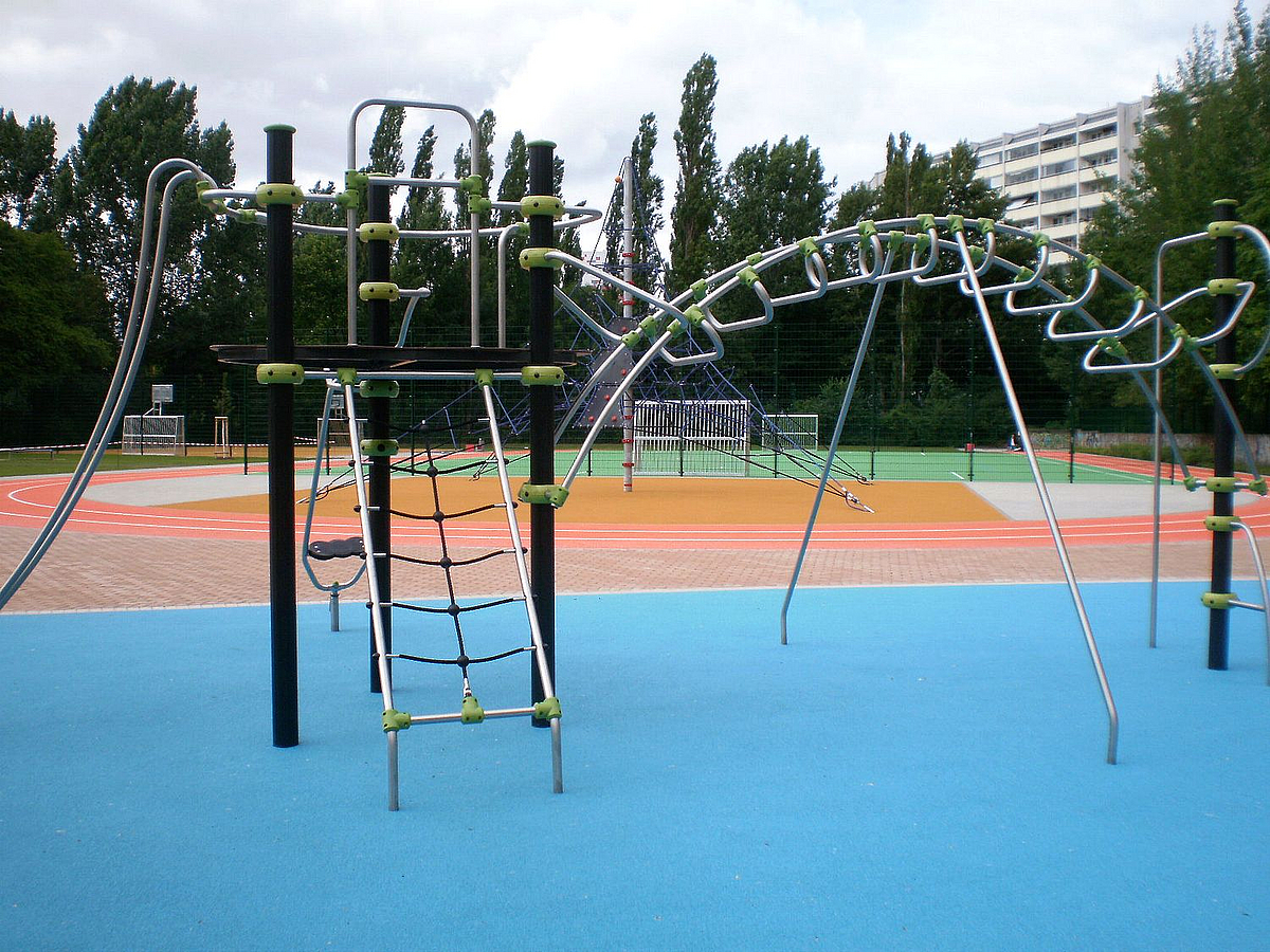 Parkour aus Stahl und Seilen auf Kunststoff, dahinter Sportflächen, Grün u. Schulgebäude