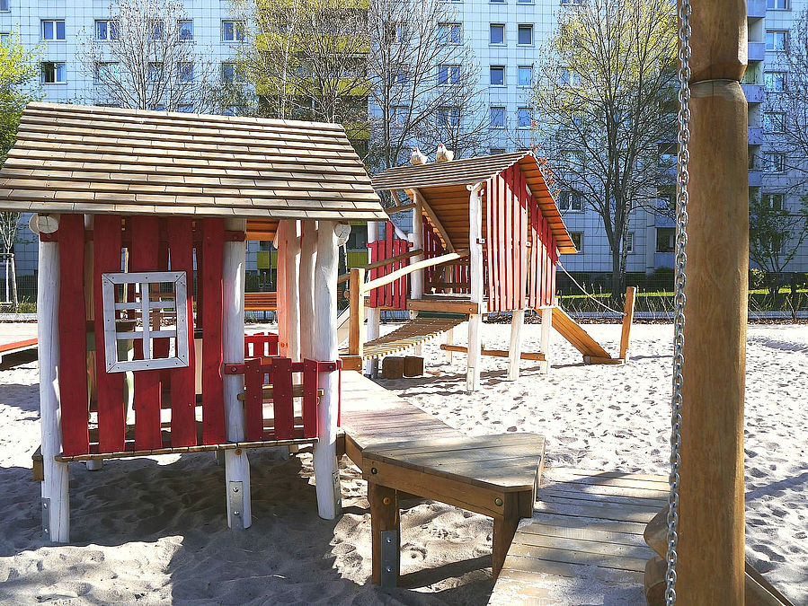 2 Spielhäuser in Rot und Weiß auf Sandfläche, dahinter Plattenbau
