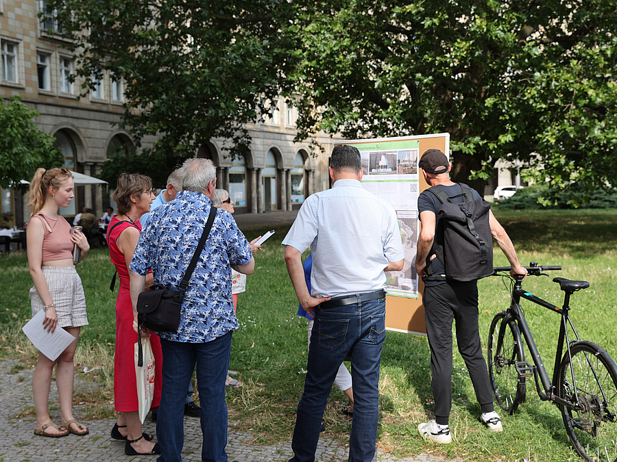Gruppe von Menschen betrachtet Tafel mit Plänen an einem grünen Platz