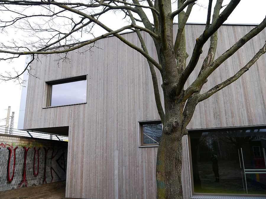 Detail Holzgebäude, dass über eine Mauer auskragt, davor kahler Baum