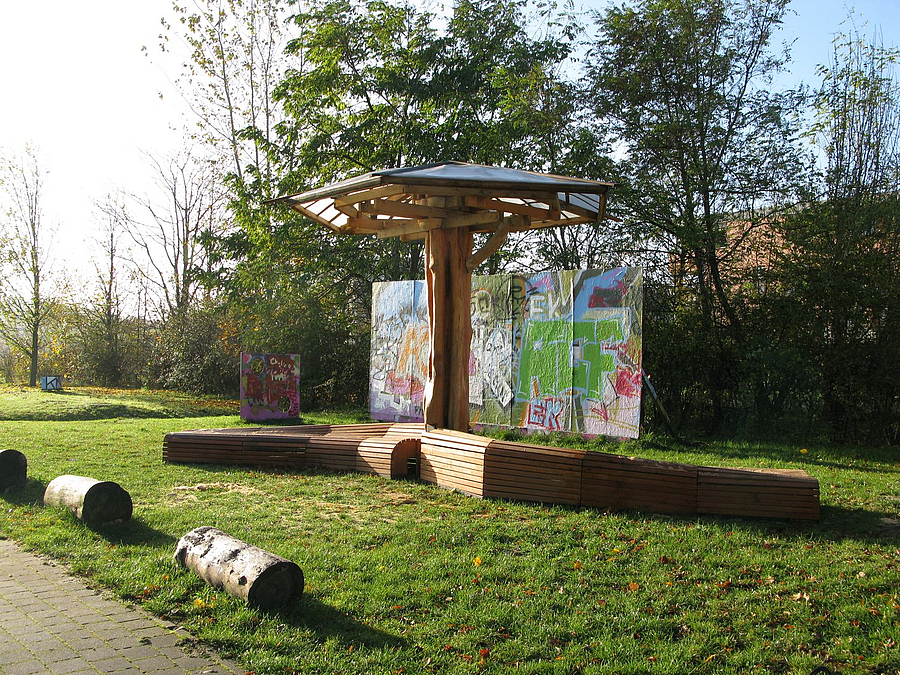 Baumartiger Pavillon mit organisch geformten Bänken aus Holzlatten auf Rasen vor Graffiti-Wand