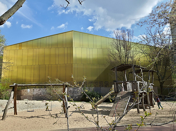 Goldene, fensterlose Fassade vor blauem Himmel, im Vordergrund Spielplatz