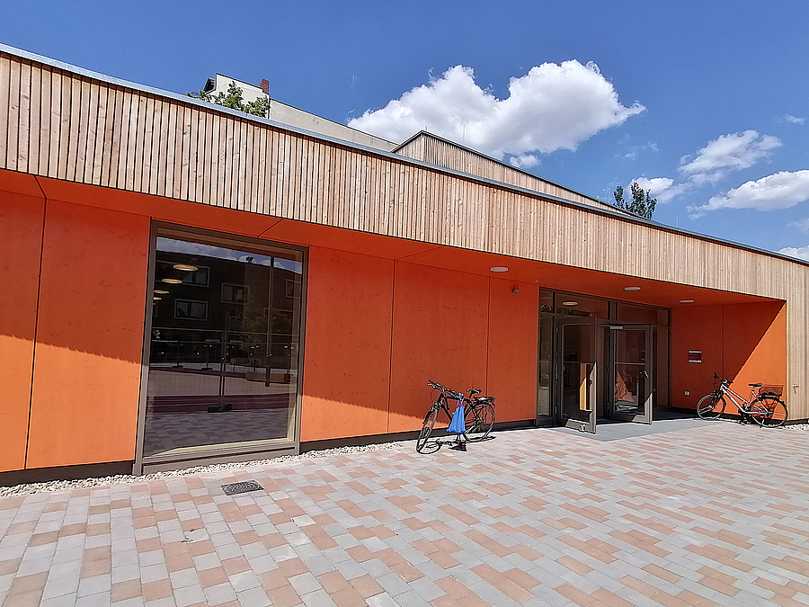 Eingeschosser, orangefarbene Front mit Eingang und Bodenfenster, oben Holzlamellenfassade