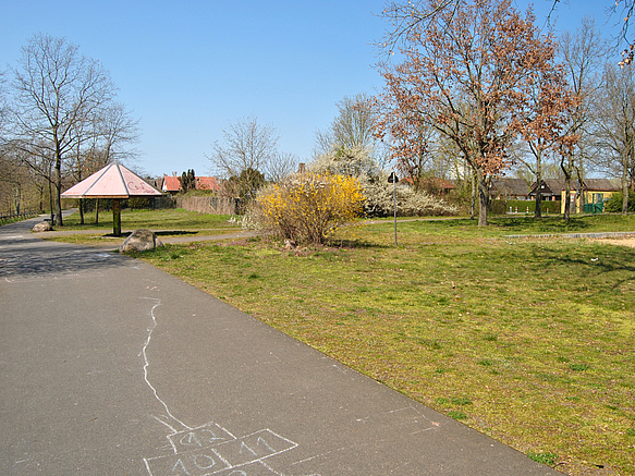 Asphaltweg mit Kreidezeichnung, Wiese, Gehölze im Frühling, pilzförmiger Pavillon