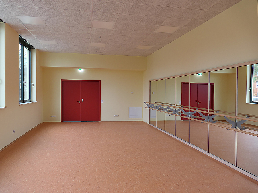 Raum mit rötlichem Boden, roter Tür, gelben Wänden und Spiegelwand mit Ballettstange