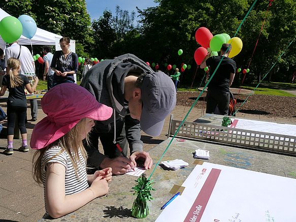 Mädchen und Mann schreiben Zettel, daneben Party-Pavillons, Luftballons