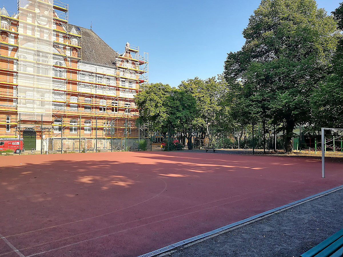 Historisches Schulgebäude eingerüstet, davor Sportplatz (Hartplatz) mit Fußballtor