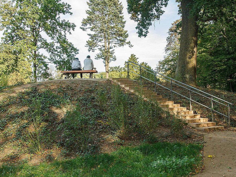Bepflanzter Hügel mit Zugangstreppe inkl. Geländer, oben 2 Menschen auf Bank