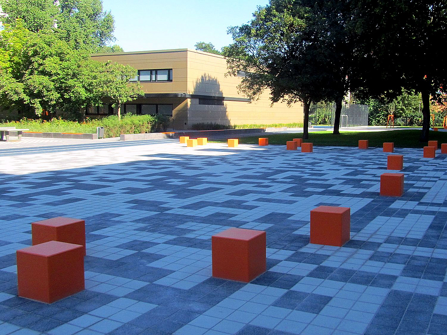 Gepflasterte Fläche im QR-Code-Muster mit orangefarbenen Sitzwürfeln, im Hintergrund Sporthalle
