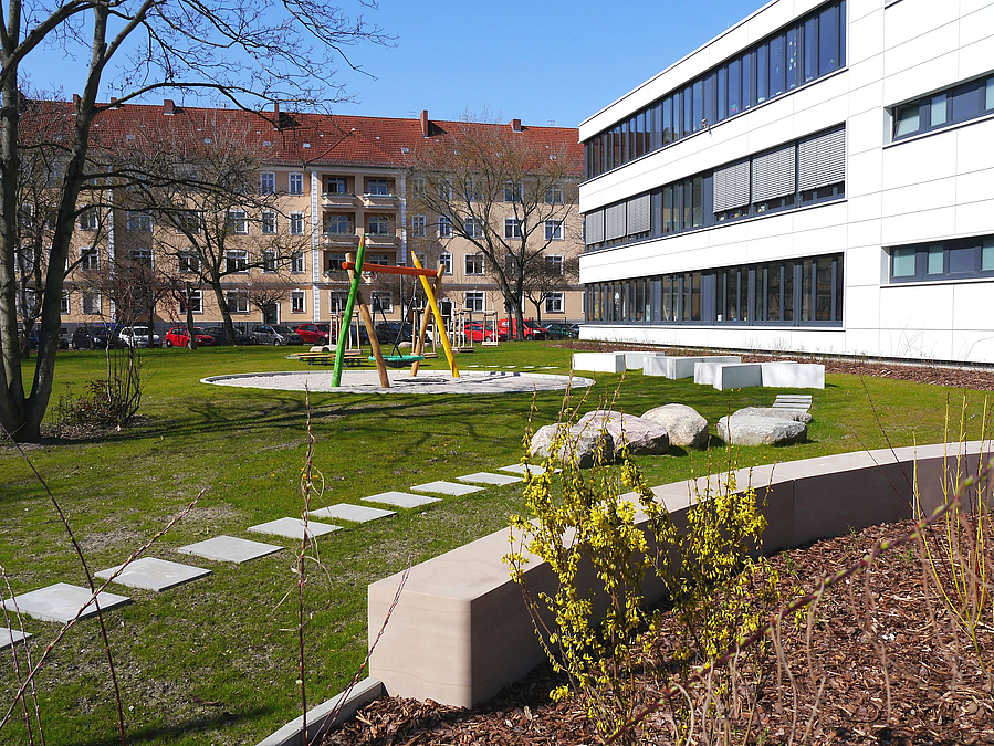 Beet mit Forsythien, Sitzmauer, Rasen, Trittplatten, weißes, dreistöckiges Schulgebäude