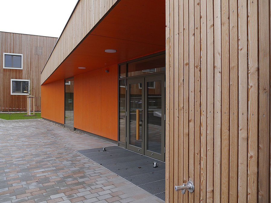 Eingang der Sporthalle als Rücksprung in der Fassade, Holzlamellen, orangefarbene Wände am Eingang