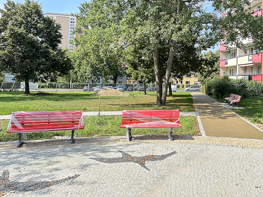 Rote Bänke mit Schutzfolie auf rundem, gepflastertem Platz mit Mosaikpflaster in Form zweier fliegender Gänse