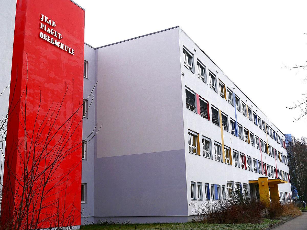 Heller viergeschossiger Schulbau mit farbig abgesetzten Fenstern, versetzter roter Verbinungsteil mit Namenszug
