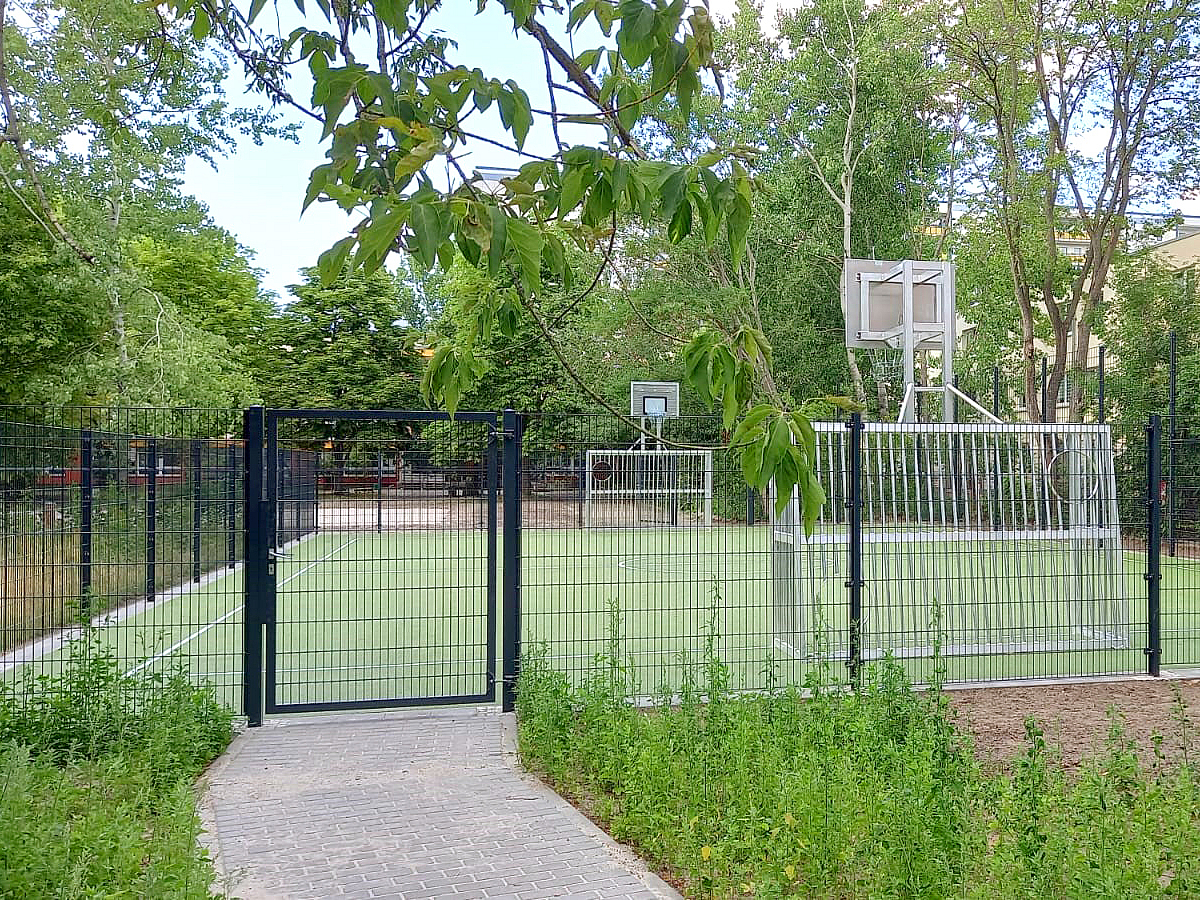 Zaun, dahinter Bolzplatz in Grün mit Basketballanlage