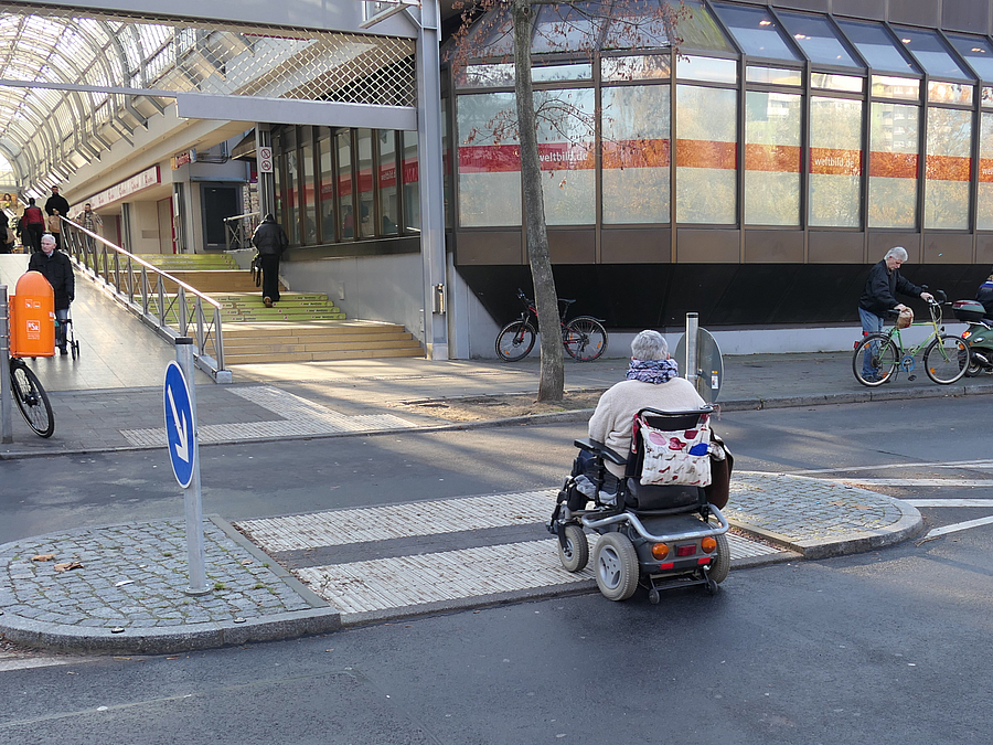 Rollstuhl fährt auf Mittelinsel mit taktilen Streifen vor einkaufszentrum
