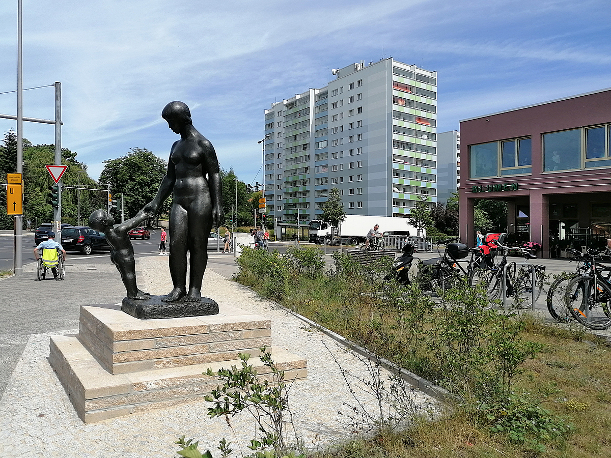 Bronzeskulptur Mutter mit Kind an der Hand, daneben Grün, Fahrräder, Einkaufszentrum, Wohnhochhäuser