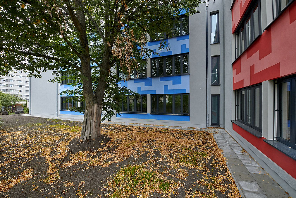Weiter Winkel zwischen Fassaden mit blauem und mit rotem Muster zwischen den Etagen, dazwischen schmale Fuge mit Fenstern, herbstlicher Baum