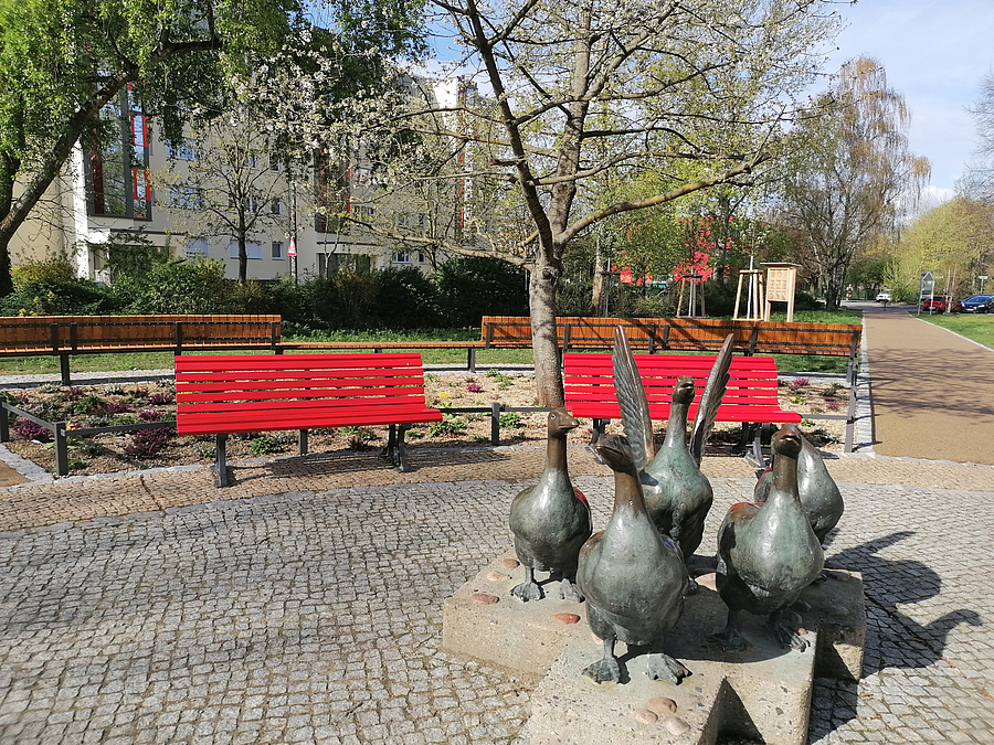 5 Gänse aus Bronze auf Sockel auf gepflastertem Platz vor roten Bänken und Bäumen