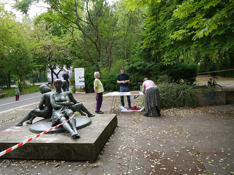 Skulptur sitzender Frauenakte vor Tisch mit 3 Menschen unter Bäumen