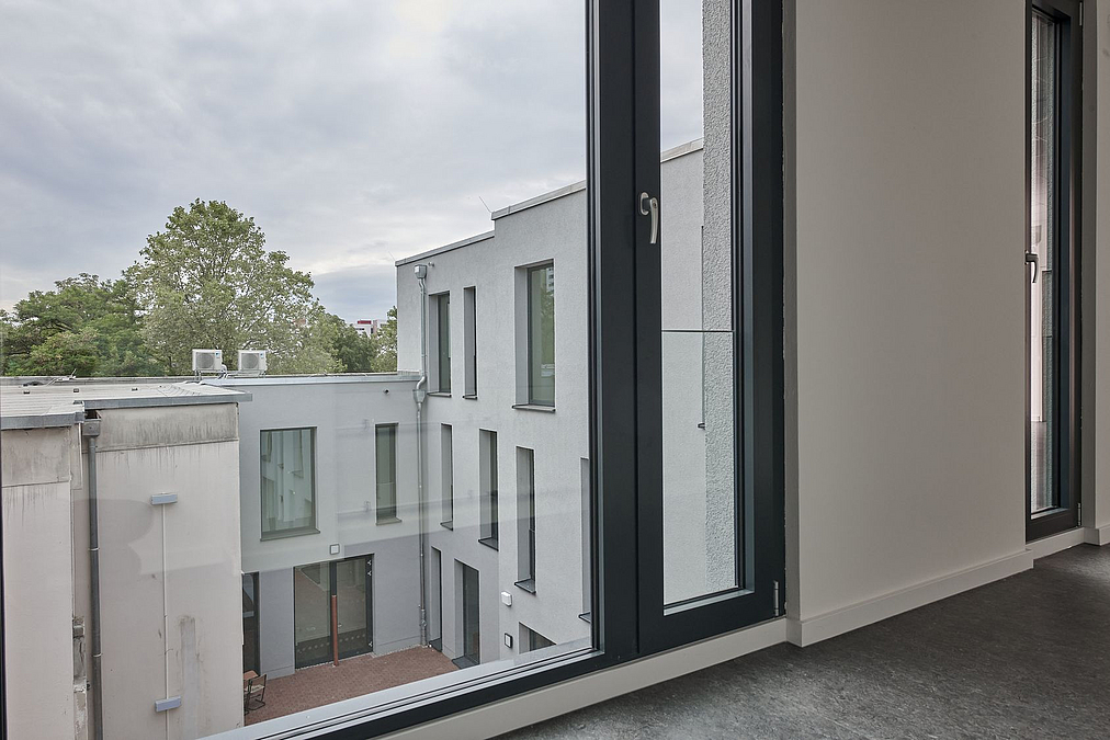 Blick aus dem Fenster auf graue dreigeschossige und zweigeschossige Fassade im rechten Winkel