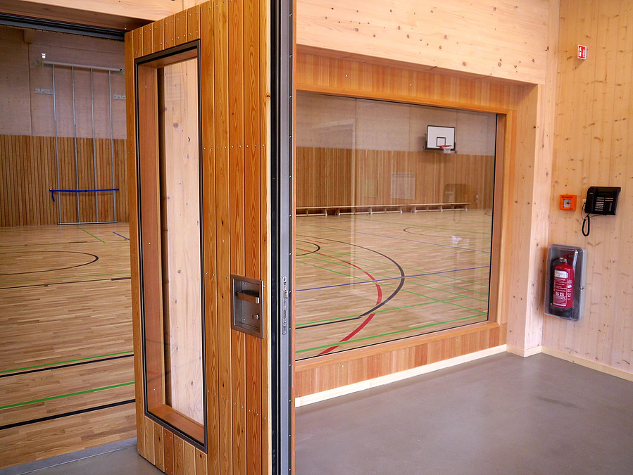 offene Holztür mit Verglasung, daneben großes Fenster zur Sporthalle