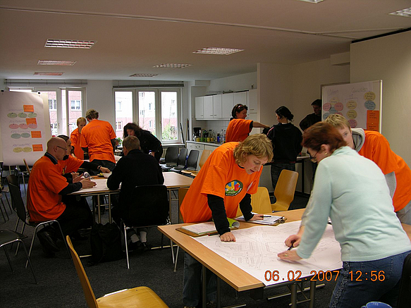 Menschen über Tische mit Plänen gebeugt, die Mehrzahl trägt orangefarbene T-Shirts mit Logo