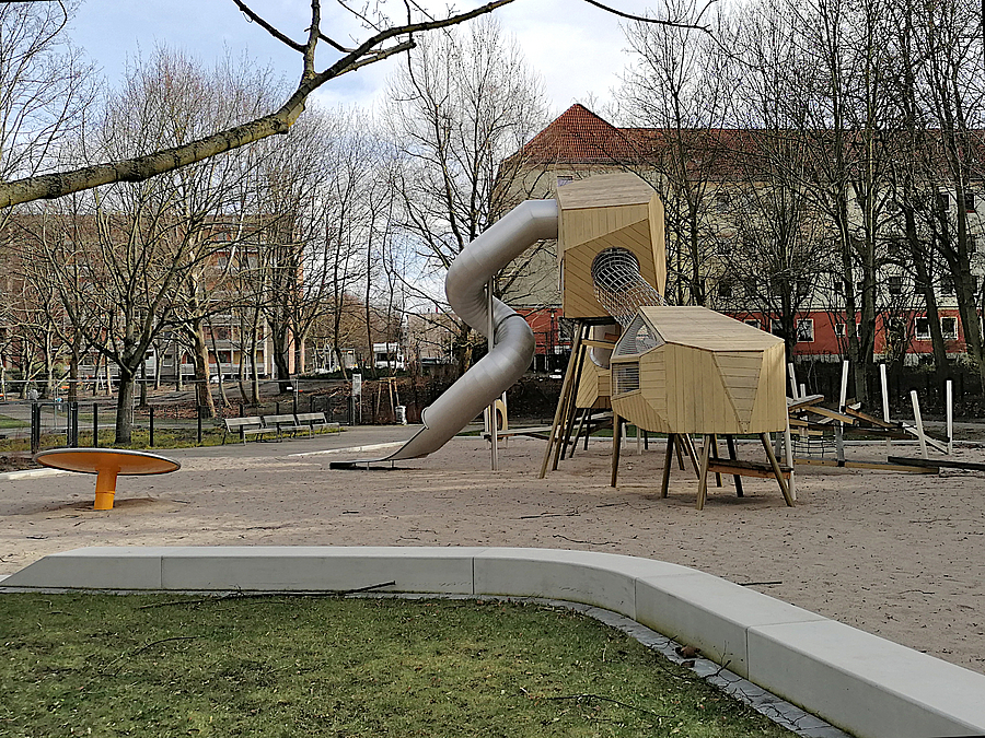 Spielplatz mit Kletter-Rutschenkombi aus geschlossenen Körpern auf Sand, davor Rasen und Sitzkante