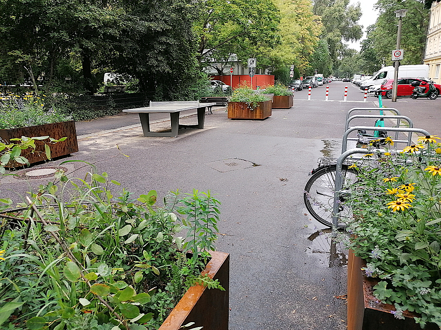 Straße mit bepflanzten Hochbeeten, Tischtennisplatte, genutzten Fahrradbügeln