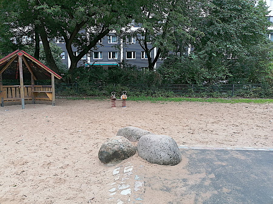 Fast leere Sandfläche, 3 kleine Findlinge, Spielhütte, dahinter Grün