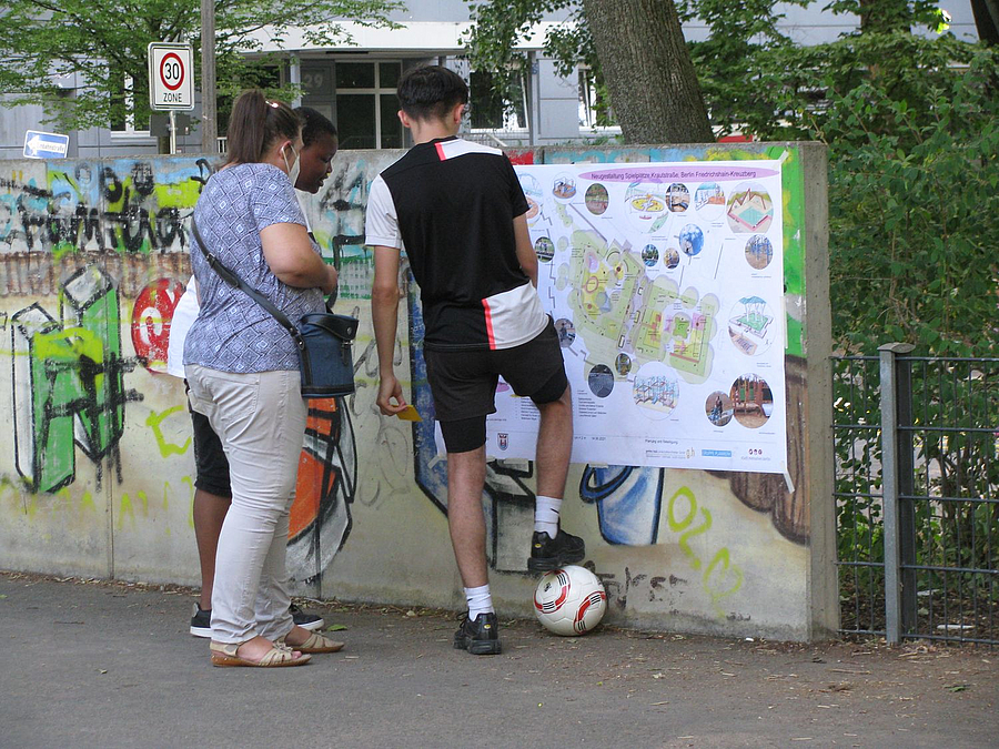 Jugendlicher mit Ball und Erwachsene sehen sich großes Plakat am Zaun an
