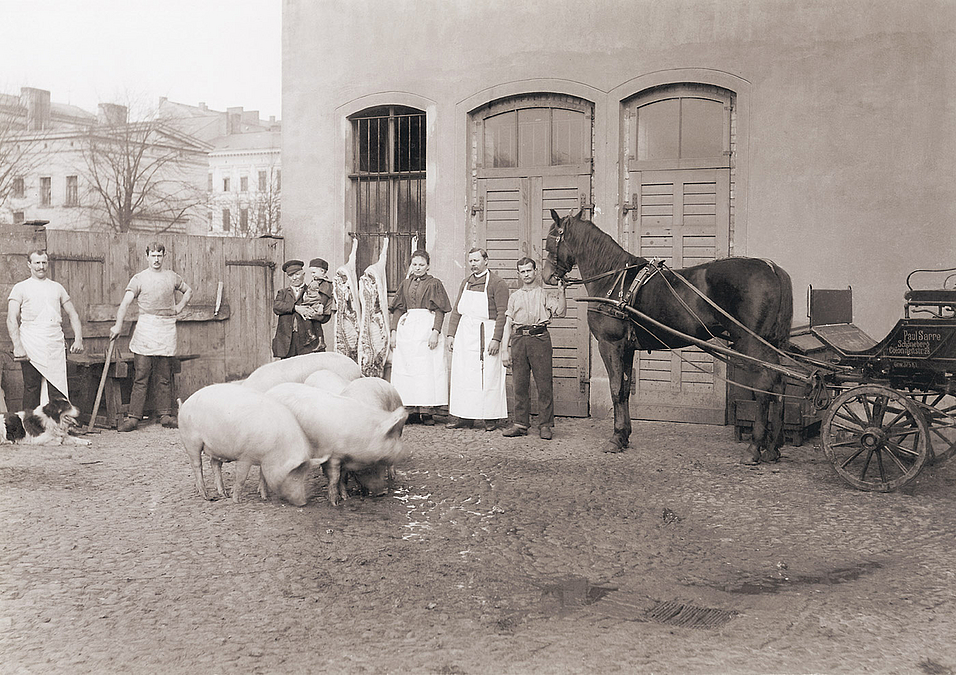 Menschen posieren in einem Hof, in der Mitte eine Gruppe Schweine, rechts ein Pferdefuhrwerk