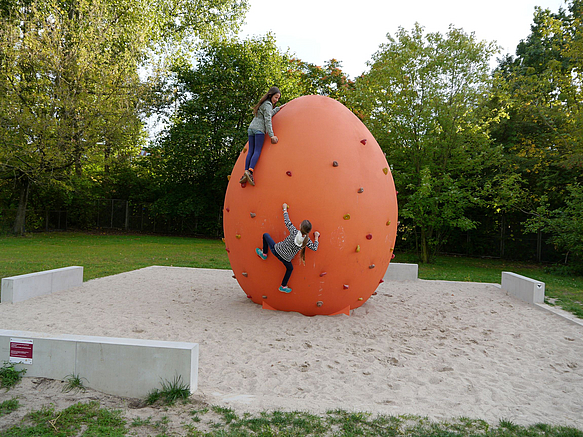 Sandfläche mit orangefarbenem Ei mit Bouldergriffen, 2 Kindern klettern