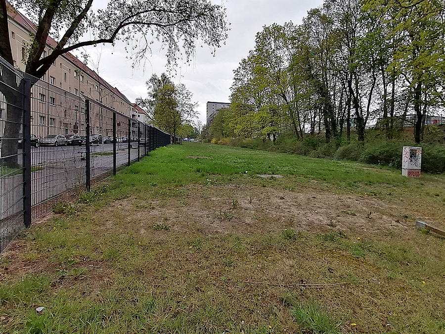 Eingezäunte Rasenfläche, rechts baumbestandener Wall, links Wohnhäuser